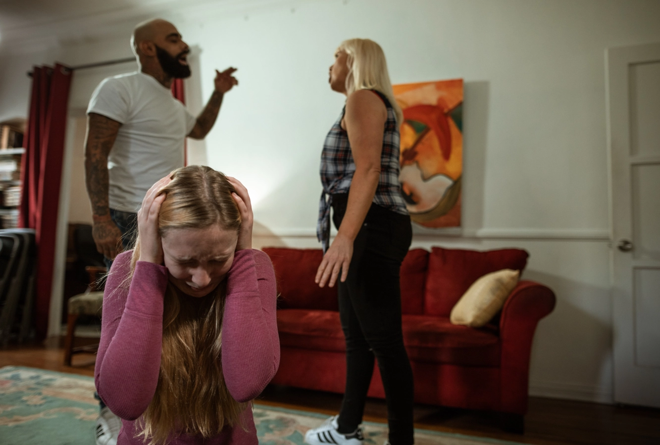 Girl with hands on head crying, while parents arguing in the background